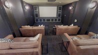This dedicated home theater is thoughtfully designed to deliver a premium cinematic experience. The room is enveloped in deep, dark-toned walls accented with a grid of decorative framed panels that frame the large central projection screen, creating a dramatic and visually striking focal point. Subtle wall sconces provide ambient lighting, contributing to the moody, immersive atmosphere while keeping the focus on the screen. The flooring features a patterned carpet that complements the room's sophisticated color palette. Seating is generously provided by a collection of oversized, plush leather sofas and chairs arranged in a tiered-style layout to ensure unobstructed sightlines from every position. Slim side tables are conveniently positioned between seats, offering practical surfaces for beverages and snacks during screenings. The layout comfortably accommodates a generous number of guests, making it equally suited for intimate viewings or larger gatherings. Every detail of this space has been curated to balance comfort and style, resulting in a high-end entertainment room that rivals a commercial theater experience.