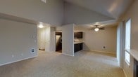 This two-floor residence is designed with an open and airy feel, highlighted by dramatically vaulted ceilings in the main living area that create a strong sense of vertical space. The living room is carpeted in a neutral tone and flows seamlessly into the kitchen, which is defined by a partial dividing wall that adds structure without sacrificing openness. The kitchen is outfitted with dark espresso-toned cabinetry, stainless steel appliances including a refrigerator, and a sink positioned along the countertop — offering a practical and cohesive workspace. A ceiling fan with a light fixture serves the kitchen and dining area, providing both air circulation and ambient lighting. Adjacent to the main living space, a hallway leads to additional rooms, with multiple doors visible — likely providing access to closets and other areas of the home. The walls throughout are finished in a soft, warm gray tone that complements the light carpeting and dark cabinetry. The overall ambiance is clean, understated, and functional, with the vaulted ceilings serving as the standout architectural feature. The second floor adds additional living space, making this a well-rounded, multi-level home suitable for a variety of lifestyles.