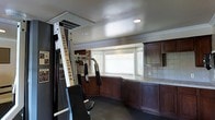 This single-floor space functions as a dedicated home gym, thoughtfully outfitted with both exercise equipment and built-in storage solutions. Dominating one side of the room is an Inflight Fitness cable machine, providing a professional-grade workout station suited for a wide range of strength exercises. The smooth, light-toned ceiling features recessed lighting and crown molding details, lending a polished, finished appearance to the room.

Along the opposite wall, rich dark-wood cabinetry with tile countertops runs the length of the space, offering ample storage and workspace. A row of wide windows positioned above the countertop allows natural light to filter into the room, brightening the interior and creating an open, airy feel. Upper cabinets flank the window arrangement on both sides, maximizing storage capacity while maintaining a cohesive, built-in aesthetic.

The flooring is dark-toned and durable, appropriate for a high-activity workout environment. The combination of functional cabinetry, natural light, and quality fitness equipment makes this space a well-organized and practical home fitness area.