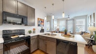 This single-floor residence offers a well-designed open layout that connects its primary living spaces seamlessly. The kitchen serves as a central hub, outfitted with taupe-toned cabinetry equipped with brushed nickel hardware, light quartz countertops, and a full suite of black appliances including a refrigerator, electric range, built-in microwave, and dishwasher. A peninsula with an undermount sink extends into the living area, while pendant lighting above adds a modern touch. Warm hardwood-style flooring runs throughout the unit, unifying the open-concept design.

The living area is bright and welcoming, benefiting from multiple large windows that allow natural light to fill the space. A ceiling fan adds to the comfortable atmosphere. The bedroom continues the same light-filled aesthetic, featuring a generous window with blinds and ample room for furnishings. The bathroom includes a wide vanity with dark cabinetry, a large mirror, and clean white countertops. A dedicated laundry room houses a full-sized washer and dryer alongside built-in shelving, offering practical storage and organizational convenience.