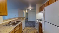 This single-floor residential space offers a practical and well-organized layout designed for comfortable everyday living. The kitchen serves as a central feature, showcasing warm honey-toned wood cabinetry that provides ample storage both above and below the counters. Granite-style laminate countertops offer generous prep space, complemented by a double-basin stainless steel sink. White appliances, including a gas range with multiple burners and a full-sized refrigerator, along with a built-in dishwasher, make the kitchen fully equipped for daily use. The flooring in the kitchen features durable, earth-toned stone-look tile that adds visual grounding to the space. A pendant light fixture overhead provides focused illumination. The kitchen flows seamlessly into a carpeted living area, which is finished in neutral tones that create a calm and inviting atmosphere. A closet with bi-fold doors is visible from the main living area, offering practical storage solutions. A hallway leads further into the unit, connecting additional rooms and maintaining an open, accessible feel throughout. Ceiling-mounted light fixtures provide consistent lighting across the space, contributing to a bright and welcoming interior overall.