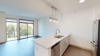 This well-appointed single-floor residence showcases a clean, contemporary design with warm wood-look flooring and crisp white walls that create a bright, welcoming atmosphere throughout. The open-concept kitchen is a standout feature, equipped with a full suite of stainless steel appliances — including a refrigerator, electric range, and over-the-range microwave — complemented by light wood cabinetry, white quartz countertops, and a classic subway tile backsplash. A central island with an undermount double sink and elegant pendant lighting serves as both a functional workspace and a natural gathering point, flowing seamlessly into the adjacent living and dining area. The living space is bathed in natural light from a large sliding glass door that opens onto a private balcony, offering treetop views that bring a sense of the outdoors inside. The bedroom is generously sized with a large window framing green outdoor views, and features a walk-in closet accessible through modern sliding barn-style doors. The bathroom is finished with a wood-toned vanity, white quartz countertop, an undermount sink, a glass-enclosed shower, and brushed nickel hardware and fixtures. An in-unit laundry area adds further convenience to this thoughtfully designed space.