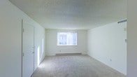 This single-floor space presents a clean, minimalist interior centered around an open main room. The room features light-toned wall-to-wall carpeting that extends throughout, contributing to a soft, understated aesthetic. Walls are finished in a crisp white, amplifying the sense of openness and brightness within the space.

A centrally positioned window fitted with vertical blinds serves as the primary source of natural light, allowing diffused daylight to fill the room. Below the window, a baseboard heating unit provides climate control, a practical and unobtrusive feature integrated into the wall. The ceiling carries a textured popcorn finish, a common architectural detail that adds subtle visual depth overhead.

A doorway is visible along one wall, providing access to an adjacent area or closet, suggesting the layout includes additional functional spaces beyond the main room. The overall ambiance is neutral and versatile, offering a blank-canvas environment well-suited for customization. The space maintains a straightforward, functional character defined by its simple finishes, natural light, and open floor plan.