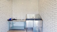 This single-floor Pet Spaw is a purpose-built pet grooming facility designed with both functionality and cleanliness in mind. The space is finished throughout with classic white subway tile walls that extend floor to ceiling, creating a bright, hygienic environment that is easy to maintain. Large-format neutral floor tiles complement the crisp tiling and contribute to the clean, professional aesthetic.

The grooming area is the centerpiece of the space, anchored by a professional-grade stainless steel pet bathing tub equipped with dedicated faucet fixtures and a wraparound splash guard. Adjacent to the tub, a stainless steel prep table provides a convenient workspace for grooming tools and supplies. A pet dryer is also available, rounding out the suite of grooming essentials.

The entry area features a dramatic floor-to-ceiling window that floods the space with abundant natural light and offers open views of the surrounding landscaped grounds and neighboring buildings. A small display shelf near the entry holds grooming products, and posted guidelines ensure the facility is used responsibly. Access is controlled via a keypad entry system. The overall ambiance is clean, practical, and welcoming for both pets and their owners.