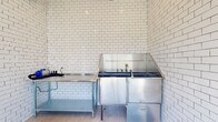 This single-floor Pet Spaw is a purpose-built pet grooming facility designed with both functionality and cleanliness in mind. The space is finished throughout with classic white subway tile walls that extend floor to ceiling, creating a bright, hygienic environment that is easy to maintain. Large-format neutral floor tiles complement the crisp tiling and contribute to the clean, professional aesthetic.

The grooming area is the centerpiece of the space, anchored by a professional-grade stainless steel pet bathing tub equipped with dedicated faucet fixtures and a wraparound splash guard. Adjacent to the tub, a stainless steel prep table provides a convenient workspace for grooming tools and supplies. A pet dryer is also available, rounding out the suite of grooming essentials.

The entry area features a dramatic floor-to-ceiling window that floods the space with abundant natural light and offers open views of the surrounding landscaped grounds and neighboring buildings. A small display shelf near the entry holds grooming products, and posted guidelines ensure the facility is used responsibly. Access is controlled via a keypad entry system. The overall ambiance is clean, practical, and welcoming for both pets and their owners.