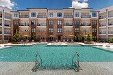 Located at 8340 Picardy Ave in Baton Rouge, Louisiana, this outdoor amenity space is a thoughtfully designed resort-style courtyard that serves as the centerpiece of a modern multi-story residential community. The space is anchored by a generously sized swimming pool with crystal-clear turquoise water, complemented by a separate spa, all framed by elegant herringbone-patterned paver decking. Surrounding the pool, an expansive sun deck is outfitted with rows of lounge chairs and shade umbrellas in a vibrant yellow-green accent, providing ample space for relaxation. A striking wood pergola adds architectural warmth to one corner of the courtyard, creating a shaded gathering area ideal for outdoor entertaining. Adjacent to the pool zone, a well-manicured lawn area and landscaped walkways connect to additional outdoor seating nodes, including umbrella-shaded dining tables nestled among mature trees and ornamental plantings. String lights overhead contribute to an inviting evening ambiance. The courtyard is enclosed by the multi-story brick-and-panel residential buildings, whose private balconies overlook the amenity area, offering sweeping views. A dedicated clubhouse structure with large glass panels opens directly onto the pool deck, seamlessly blending indoor and outdoor living. The overall atmosphere is polished, vibrant, and resort-inspired.