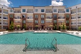Located at 8340 Picardy Ave in Baton Rouge, Louisiana, this outdoor amenity space is a thoughtfully designed resort-style courtyard that serves as the centerpiece of a modern multi-story residential community. The space is anchored by a generously sized swimming pool with crystal-clear turquoise water, complemented by a separate spa, all framed by elegant herringbone-patterned paver decking. Surrounding the pool, an expansive sun deck is outfitted with rows of lounge chairs and shade umbrellas in a vibrant yellow-green accent, providing ample space for relaxation. A striking wood pergola adds architectural warmth to one corner of the courtyard, creating a shaded gathering area ideal for outdoor entertaining. Adjacent to the pool zone, a well-manicured lawn area and landscaped walkways connect to additional outdoor seating nodes, including umbrella-shaded dining tables nestled among mature trees and ornamental plantings. String lights overhead contribute to an inviting evening ambiance. The courtyard is enclosed by the multi-story brick-and-panel residential buildings, whose private balconies overlook the amenity area, offering sweeping views. A dedicated clubhouse structure with large glass panels opens directly onto the pool deck, seamlessly blending indoor and outdoor living. The overall atmosphere is polished, vibrant, and resort-inspired.