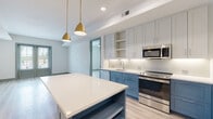 This thoughtfully designed single-floor residence showcases a contemporary aesthetic with a cohesive color palette and high-quality finishes throughout. The open-concept kitchen serves as the heart of the space, featuring a striking two-tone cabinetry design that pairs crisp white upper cabinets with bold blue lower cabinets. A spacious central island with a pristine white countertop provides generous prep and dining space, illuminated by a pair of elegant gold pendant lights. Stainless steel appliances, including a French door refrigerator, range, dishwasher, and built-in microwave, complete the well-appointed kitchen. White subway tile backsplash and recessed lighting add a clean, polished touch. Light-toned hardwood-style flooring flows seamlessly through the living areas and bedrooms, creating a warm and unified feel. The bedrooms are bright and airy, benefiting from large windows with outdoor views and ceiling fans for added comfort. French doors in the main living area enhance the sense of openness and invite natural light. The bathroom features warm terracotta hexagonal floor tiles, a dual-sink vanity with blue cabinetry, backlit mirrors, and a glass-enclosed walk-in shower with classic white subway tile, delivering a spa-like atmosphere.