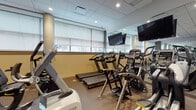 Located at 695 Atlantic St in Stamford, Connecticut, this well-appointed fitness center offers a comprehensive workout environment designed to accommodate a wide range of exercise routines. The space is laid out on a single floor and is outfitted with an extensive selection of cardio machines, including treadmills, ellipticals, stationary bikes, and adaptive motion trainers. A dedicated strength-training area features a multi-station cable machine, a full dumbbell rack, weight benches, and additional resistance equipment. The flooring throughout consists of durable, textured rubber material suited for high-activity use. Large windows fitted with roller shades line one wall, allowing natural light to filter into the space while maintaining privacy and glare control. Full-length mirrors are strategically positioned along the walls, amplifying the sense of space and supporting proper form during workouts. Wall-mounted flat-screen televisions are placed throughout for entertainment during cardio sessions. The drop ceiling incorporates recessed lighting that ensures even, bright illumination across the entire floor. Glass double doors provide clear sightlines and easy access into the facility. The overall design is modern and functional, with a neutral color palette of gray and tan tones contributing to a focused, professional training atmosphere.