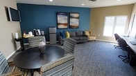 This inviting single-floor lounge is thoughtfully designed to provide a comfortable and functional gathering space. The room is anchored by a bold teal feature wall adorned with two large abstract paintings, adding a striking visual focal point that complements the warm neutral tones of the surrounding walls. Plush seating options include a gray sofa accented with blue and yellow throw pillows, a tan accent chair, and a pair of patterned armchairs positioned around a round dark-topped table — ideal for casual conversation or a quiet retreat. A compact refreshment station in the corner features a coffee maker and a mini refrigerator, offering added convenience for occupants. The space is finished with a patterned carpet that ties the color palette together, recessed lighting overhead, and a window that allows natural light to filter in. The overall ambiance is modern yet comfortable, striking a balance between professional presentation and relaxed hospitality, making it well-suited as a community lounge, waiting area, or shared amenity space.
