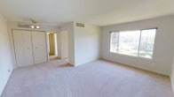 This single-floor residential space offers a clean, functional layout with a neutral color palette that creates a bright and welcoming atmosphere throughout. The main room is generously proportioned, featuring soft wall-to-wall carpeting that adds warmth underfoot and a crisp white ceiling that enhances the sense of openness. A large window allows abundant natural light to fill the space while offering a pleasant view of the greenery outside, further contributing to the airy feel of the interior. A built-in closet with sliding doors provides convenient and ample storage, making the room both practical and organized. A ceiling-mounted light fixture adds supplementary illumination. The HVAC vent integrated into the wall ensures year-round climate comfort. A hallway visible from the main room connects to additional areas of the space, all finished with consistent light-toned carpeting and white walls that maintain a cohesive, clean aesthetic. The overall ambiance is calm and understated, offering a neutral canvas well-suited for a variety of uses and personal styling preferences. The single-floor layout provides ease of access and seamless flow between areas.