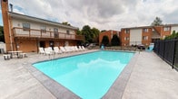 Aura is a well-appointed community space designed to offer residents a comfortable and functional environment. The centerpiece of this single-floor property is the expansive outdoor pool area, which serves as an inviting retreat for relaxation and recreation. The pool itself is generously sized, filled with clear, blue water, and bordered by a clean, wide concrete deck. A row of white lounge chairs lines one side of the pool, providing ample space for sunbathing and leisure. The pool area is enclosed by a black iron fence, ensuring safety and privacy, while mature trees and landscaped greenery in the background lend a natural, serene atmosphere. An elevated wooden deck adjoins the neighboring building, adding an additional vantage point overlooking the pool. Beyond the outdoor amenity space, the property includes interior utility areas and a modern office or business center, visible through a glass-paneled door. The office space is thoughtfully equipped with a computer workstation and tasteful wall art, offering a quiet, professional setting for residents' use. The overall aesthetic of Aura balances practical amenities with comfortable communal spaces, making it a well-rounded destination for both everyday living and leisure.