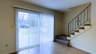 This three-floor space offers a thoughtfully designed layout that makes effective use of its vertical footprint. The main level immediately draws attention to a large sliding glass door dressed with full-length vertical blinds, flooding the area with natural light and providing views of the surrounding outdoor greenery and street. The flooring on this level features neutral-toned tile, lending a clean and practical foundation to the space.

A split-level transition is evident, with a carpeted platform rising a few steps up from the main floor, creating a subtle separation of zones within the open living area. This raised section connects to a staircase finished in matching carpet, framed by a sleek metal railing that adds a modest contemporary touch. The staircase leads to the upper floors, suggesting additional living or sleeping areas above.

The walls throughout are finished in a warm neutral tone, contributing to a calm and understated ambiance. Recessed floor vents indicate a central heating system integrated into the structure. Overall, the space presents a functional and straightforward multi-level layout with a practical, livable character.