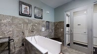 This single-floor Pet Spa is purpose-built to provide a premium grooming experience, combining functional design with refined aesthetic details. The space is finished with large-format natural stone tiles in rich earth tones, covering both the walls and floors to create a cohesive, upscale environment. A deep, freestanding-style soaking tub takes center stage, clad in matching stone tile and equipped with a wall-mounted spray fixture for convenient bathing. Adjacent to the tub is a separate enclosed shower area, framed with sleek glass and metal paneling, providing an additional bathing option. The walls are painted in a soft, calming blue-gray tone that complements the warm stone surfaces and contributes to a serene atmosphere. Playful framed artwork depicting animals adorns the walls, adding a lighthearted and welcoming character to the space. Thoughtful details such as mounted informational panels and accessible fixtures reflect a practical, user-friendly layout. Overall, the Pet Spa balances elegance with functionality, offering a clean, well-appointed environment designed to make the grooming process comfortable for pets and convenient for their owners.