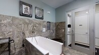 This single-floor Pet Spa is purpose-built to provide a premium grooming experience, combining functional design with refined aesthetic details. The space is finished with large-format natural stone tiles in rich earth tones, covering both the walls and floors to create a cohesive, upscale environment. A deep, freestanding-style soaking tub takes center stage, clad in matching stone tile and equipped with a wall-mounted spray fixture for convenient bathing. Adjacent to the tub is a separate enclosed shower area, framed with sleek glass and metal paneling, providing an additional bathing option. The walls are painted in a soft, calming blue-gray tone that complements the warm stone surfaces and contributes to a serene atmosphere. Playful framed artwork depicting animals adorns the walls, adding a lighthearted and welcoming character to the space. Thoughtful details such as mounted informational panels and accessible fixtures reflect a practical, user-friendly layout. Overall, the Pet Spa balances elegance with functionality, offering a clean, well-appointed environment designed to make the grooming process comfortable for pets and convenient for their owners.