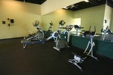 Located at 2100 Westminster Ln in Virginia Beach, Virginia, this single-floor commercial fitness facility offers a well-organized and functional workout environment. The space features an open-concept layout with durable rubber flooring throughout, providing a safe and practical surface for a wide range of exercise activities.

The main workout area is generously proportioned, accommodating an array of cardio equipment including treadmills, ellipticals, and rowing machines, all arranged to maximize floor space and user accessibility. Large wall-mounted mirrors line multiple sections of the facility, enhancing the sense of openness and allowing users to monitor their form during workouts. A dedicated strength training zone features a multi-function cable machine, free weights, and benches, offering versatile resistance training options.

The facility benefits from an exposed black ceiling with industrial-style structural elements, ceiling fans, and fluorescent lighting that collectively create a utilitarian, high-energy atmosphere. Large storefront windows along one wall allow natural light to fill the space and provide views of the surrounding exterior. A tiled entryway leads into the main floor area, and safety equipment such as a fire extinguisher is visibly positioned near the entrance. The overall design prioritizes functionality, making it well-suited for personal training, group fitness, or general gym use.
