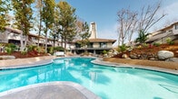 Located at 8211 San Angelo Dr in Huntington Beach, California, this single-level outdoor amenity space offers a well-appointed recreational environment designed for both relaxation and community enjoyment. At the heart of the space is a generously sized freeform swimming pool with crystal-clear turquoise water, complemented by a separate circular spa, both finished with classic tile detailing and surrounded by a smooth concrete pool deck. A distinctive clubhouse building with a striking tower element and Spanish-influenced tile roofing serves as an architectural focal point, lending the area a resort-like character. The grounds have been thoughtfully refreshed with newly installed landscaping, featuring an array of ornamental grasses, bromeliads, palms, and tropical accent plants set against a backdrop of mature pine, eucalyptus, and cypress trees. Boulder accents and layered terraced hillsides add natural texture throughout the space. Winding concrete pathways with decorative lighting guide residents through the lushly planted grounds, connecting key areas of the site. Lounge seating is positioned poolside, and the open-air setting benefits from the abundant Southern California sunshine. The overall ambiance is tranquil and resort-inspired, reflecting a well-maintained community space ideally suited to the mild coastal climate of Huntington Beach.