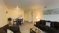 This single-floor residence offers a thoughtfully arranged open-concept layout that seamlessly connects the living, dining, and kitchen areas. The living room is a comfortable and welcoming space, featuring wall-to-wall carpeting that adds warmth underfoot. It is furnished with ample seating and accented by soft, ambient lighting that creates a cozy atmosphere throughout.

Adjacent to the living area, the dining space accommodates a full dining set and benefits from its proximity to the kitchen, which is accessible via a convenient pass-through window — a practical architectural detail that enhances both functionality and the open feel of the shared space. The kitchen pass-through also allows natural sightlines across the main living area, making the space feel more connected and expansive.

The neutral color palette — featuring soft white walls and light-toned carpeting — provides a clean, versatile backdrop that suits a variety of interior styles. Recessed HVAC vents are integrated into the ceiling and walls, ensuring year-round climate comfort. A hallway visible from the main living area leads to additional rooms, suggesting a well-organized and efficient floor plan. Overall, the space presents a functional and comfortable single-floor layout ideal for everyday living.