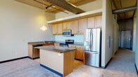 This 2-bedroom, 2-bathroom urban loft offers a distinctive industrial-chic aesthetic defined by exposed cylindrical ductwork, track lighting, and soaring ceilings that lend the space an open, airy feel. The open-concept kitchen is a functional centerpiece, featuring warm maple cabinetry, dark granite countertops, a central island, and a full suite of stainless steel appliances including a French door refrigerator, electric range with microwave, and dishwasher. A pendant light hangs above the island, adding a stylish touch to the space. The kitchen flows seamlessly into the carpeted living area, creating an inviting, cohesive layout. Both bedrooms are generously proportioned and carpeted, with large multi-pane windows that frame impressive city skyline views, flooding the rooms with abundant natural light. The primary bathroom is well-appointed with a dual-sink vanity topped with granite countertops, a walk-in glass shower, tile flooring, and a large mirror with vanity lighting. The second bathroom offers a granite-topped vanity, a glass-enclosed tub and shower combination, and matching tile flooring. Each closet provides practical storage. The overall aesthetic balances urban industrial character with comfortable residential finishes throughout.