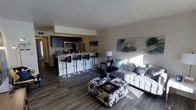 This single-floor space showcases a thoughtfully designed open-concept layout that integrates the kitchen and living area into one cohesive, functional environment. The kitchen is outfitted with dark cabinetry, stainless steel appliances including a microwave, refrigerator, and range, and is anchored by a raised breakfast bar with sleek adjustable bar stools — ideal for casual dining or entertaining. Rich, dark hardwood-style flooring runs throughout the space, providing a warm contrast to the neutral-toned walls and creating a sense of continuity between areas. The living area is generously proportioned, bathed in natural light from large windows, and features a tufted light-colored sofa accented with decorative pillows. A bold, patterned ottoman serves as a striking centerpiece, doubling as a coffee table. Abstract artwork adorns the walls, adding a contemporary artistic touch to the overall aesthetic. The smooth white ceilings and clean lines throughout contribute to a modern, uncluttered ambiance. This well-appointed space balances comfort and style, making it both practical for everyday living and visually engaging for guests.