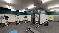 This fitness center at 1435-1437 SE Alder St in Portland, OR, is a thoughtfully designed single-floor space offering a comprehensive range of exercise equipment and amenities. The interior features a distinctive two-tone color scheme, with deep teal upper walls contrasting against crisp white lower walls, creating a modern and energizing atmosphere. Rubber flooring throughout provides durability and cushioning underfoot.

The space is divided into two connected areas. The cardio zone houses Matrix-brand treadmills, upright and recumbent stationary bikes, and an elliptical trainer, alongside a water bottle filling station and drinking fountain for convenient hydration. Narrow clerestory windows along the upper wall allow natural light to supplement the overhead fluorescent lighting.

The strength and flexibility area features a Matrix functional trainer cable system with TRX suspension attachments, a dumbbell rack with varying weights, and adjustable workout benches. Floor-to-ceiling mirrors line the walls, visually expanding the space and allowing users to monitor their form. A wall-mounted flat-screen TV, exercise mats, stability balls, and potted plants complete the space, contributing to a functional yet inviting workout environment.