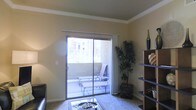 This single-floor space offers a well-appointed living area designed for both comfort and functionality. The room is finished with crown molding along the ceiling, adding a refined architectural detail that elevates the overall aesthetic. Warm, neutral-toned walls provide a versatile backdrop that complements a variety of décor styles.

A prominent feature of the living area is the large sliding glass door, which serves as both a source of abundant natural light and a direct access point to a private outdoor balcony. The balcony is outfitted with seating furniture, making it a functional extension of the interior living space. Views of surrounding greenery and neighboring structures are visible through the glass, contributing to a connected, open feel.

Inside, the living space accommodates a glass-top coffee table set atop a plush area rug, alongside a sleek dark-toned shelving unit positioned near the entrance. The shelving provides both display and storage functionality, with decorative accents adding visual interest. A tall indoor plant near the balcony door introduces a natural element, enhancing the room's inviting ambiance. The overall layout is efficient and thoughtfully arranged to maximize the sense of openness throughout.