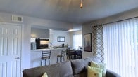 Located at 7680 West Highway 98 in Pensacola, Florida, this single-story residence offers a well-organized and functional layout across several distinct living areas. The open-concept design connects the living room, kitchen, and dining area into a cohesive, sociable space. The living room features a textured ceiling with a ceiling fan and large windows dressed with patterned curtains that allow natural light to fill the room. The kitchen is positioned behind a raised breakfast bar with barstool seating, providing a casual dining option alongside the dedicated dining area. Dark appliances, including a full-sized refrigerator and range, anchor the kitchen, which also features white cabinetry and ample counter space. The dining area accommodates a full table and chair set, making it ideal for everyday meals or entertaining. A hallway connects the common areas to the private quarters, which include at least two bedrooms. One bedroom is notably spacious and features a dresser and room for a full bedroom suite. The overall color palette throughout the home is neutral and calming, with gray tones and white walls contributing to a clean, modern aesthetic. The floor plan is efficient and practical, making excellent use of the available square footage across its single level.