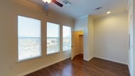 This single-story space presents a well-appointed room characterized by rich, dark hardwood flooring that extends throughout the interior, creating a warm and grounded aesthetic. Crown molding lines the perimeter of the ceiling, adding a refined architectural detail that elevates the overall finish of the space. A ceiling fan is centrally mounted, providing both functional air circulation and a subtle design element overhead.

Two generously sized windows dominate one wall, fitted with horizontal blinds and allowing an abundance of natural light to fill the room. The windows offer views of surrounding foliage and what appears to be a scenic outdoor landscape, enhancing the sense of openness within the space. The walls are finished in a neutral, warm beige tone that complements the flooring and contributes to a clean, inviting atmosphere.

An open door reveals a closet with a built-in hanging rod, providing practical storage directly within the room. The closet interior is finished with the same warm tones as the main space. Dual HVAC vents are visible in the ceiling, indicating a central climate control system. Overall, the space balances functional amenities with tasteful architectural detailing.