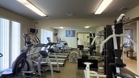 This fitness center is a single-floor facility designed to support a comprehensive workout experience. The space is outfitted with a solid lineup of cardio equipment, including multiple treadmills and an elliptical trainer, all arranged to allow comfortable movement between machines. Strength training is well represented with a multi-station cable and weight machine setup, along with a weight bench and a rack of free weights, catering to a range of exercise routines.

Large mirrors line the walls, visually expanding the space and allowing users to monitor their form during workouts. Bright fluorescent overhead lighting ensures the room is well-lit throughout. A floor fan provides additional air circulation, contributing to a comfortable training environment. Motivational fitness posters are displayed on the walls, adding an energetic ambiance to the space.

Natural light filters in through a window on one side of the room, offering a refreshing connection to the outside. The carpeted flooring provides a practical and cushioned surface underfoot. Overall, the fitness center is thoughtfully arranged to maximize usable floor space while offering a diverse range of equipment suitable for both cardio and resistance training.