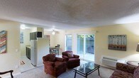 This single-floor space features an open-concept layout that connects the living area, dining space, and kitchen in one cohesive flow. The living room is carpeted and comfortably sized, furnished with upholstered armchairs and a glass-top coffee table. A textured ceiling with a ceiling fan overhead adds functional character to the room. Large sliding glass doors along one wall flood the space with natural light and provide direct access to a private outdoor patio enclosed by a fence, offering a quiet exterior retreat with visible greenery beyond.

Adjacent to the living area, a dining nook accommodates a small table and chairs, positioned near a window that further contributes to the bright, airy atmosphere of the space. The kitchen is partially visible from the main living area, separated by a half-wall partition. Standard appliances, including a refrigerator, can be seen, indicating a practical and functional kitchen setup. A wall-mounted HVAC unit provides climate control. The warm yellow-toned walls throughout contribute to a welcoming and casual ambiance, making the space feel inviting and easy to navigate.