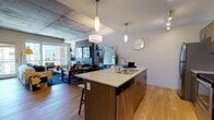 This single-floor urban residence showcases a thoughtfully designed open-concept layout that seamlessly connects the kitchen, dining, and living areas. The kitchen is a standout feature, equipped with stainless steel appliances including a refrigerator, range, and microwave, complemented by rich walnut-toned cabinetry, light quartz countertops, and a central island with a double sink and bar seating. Track lighting and pendant lights illuminate the space with a warm, modern glow, while light hardwood flooring runs throughout the main living areas.

The living room exudes a contemporary aesthetic, highlighted by exposed concrete ceilings that lend an industrial character to the space. Large windows fill the room with natural light and frame views of the surrounding cityscape. A boldly accented feature wall anchors the living area, creating a stylish focal point.

The bedroom is a serene retreat, carpeted for comfort and bathed in natural light from expansive windows overlooking the city. A ceiling fan adds functionality, and the room offers ample closet space accessed via a carpeted hallway. The bathroom is clean and well-finished, featuring large-format tile flooring, a full soaking tub with classic white subway tile surround, and warm wood vanity cabinetry.