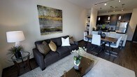 This single-floor space showcases a thoughtfully designed open-concept layout that seamlessly connects the living, dining, and kitchen areas into one cohesive environment. The living area is anchored by a deep charcoal sofa adorned with decorative accent pillows in gold and earth tones, complemented by a sleek glass-top coffee table resting on a plush area rug. A striking large-scale abstract artwork in grey and gold tones serves as a focal point on the living room wall, adding a bold artistic element to the space. Dark hardwood flooring flows continuously throughout, lending warmth and a sense of sophistication. The dining area features a modern wood-top table paired with contemporary white and chrome chairs, creating an inviting space for meals and gatherings. A set of pendant lights hangs above, providing both task lighting and a design accent. Adjacent to the dining space, the kitchen is outfitted with dark cabinetry and open shelving, offering both style and practicality. Track lighting runs along the ceiling, ensuring the space is well-lit throughout. The overall ambiance is modern and refined, with a cohesive neutral color palette punctuated by gold and dark tones that create an atmosphere of understated elegance.