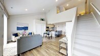 This two-storey space showcases a thoughtfully designed open-concept main level where the living, dining, and kitchen areas flow seamlessly into one another. The soaring double-height ceilings in the living area create an immediate sense of spaciousness and airiness, drawing the eye upward toward the loft level above, which is accessible via a staircase positioned along the right side of the main floor.

The living area is anchored by smooth, neutral-toned walls and recessed lighting, complementing the warm wood-look flooring that runs throughout. A cozy seating arrangement with a grey sofa and decorative accents adds a welcoming touch. Adjacent to this, the dining area accommodates a full-sized table and chairs, transitioning naturally into the kitchen.

The kitchen features light wood cabinetry, stainless steel appliances, and a large island with bar-style seating — ideal for casual dining or entertaining. Pendant lighting above the island adds a modern finishing touch. The loft level, partially visible from below, offers additional functional space. Overall, the design balances clean lines, neutral tones, and smart vertical use of space.