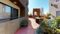 Located in the heart of downtown San Diego, this thoughtfully designed outdoor terrace offers a compelling blend of urban energy and relaxed California living. The space features a distinctive terracotta-toned concrete floor that anchors the entire area, providing a warm, sun-drenched atmosphere throughout the day.

The terrace is richly landscaped with a diverse mix of drought-tolerant and subtropical plants, including succulents, ornamental grasses, flowering shrubs, and agaves, all arranged in generous planters that create a lush, garden-like environment. A decorative archway structure with a Southwestern-influenced design adds architectural character and serves as a striking focal point toward one end of the terrace.

Comfortable outdoor seating areas featuring wicker lounge chairs with plush cushions are strategically positioned to take full advantage of the surroundings. Views extend to a well-maintained courtyard below, complete with a turf lawn and recreational game setup, as well as sweeping sightlines toward the downtown San Diego skyline, where high-rise towers rise prominently against the clear blue sky.

The multi-story building facade, finished in textured stucco with recessed balconies, frames the space on multiple sides, reinforcing a sense of enclosure and privacy. Abundant natural light fills the terrace year-round, making it an inviting outdoor retreat in the heart of the city.