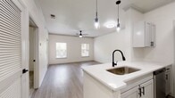 This contemporary apartment showcases a thoughtfully designed open floor plan. The main living area boasts hardwood flooring, high ceilings with modern light fixtures, and large windows that flood the space with natural light. The kitchen features white cabinetry, quartz countertops, and stainless steel appliances, seamlessly integrating with the living space. The bathroom offers a clean, monochromatic design with a striking black and white tiled accent. Bedrooms are carpeted for comfort, with ample natural light and ceiling fans. Throughout, the apartment maintains a crisp, neutral color palette, creating a bright and airy atmosphere perfect for modern living.