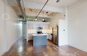 Located at 2031–2055 Ovaltine Ct in Villa Park, IL, this 670-square-foot, one-bedroom, one-bathroom unit offers a distinctive loft-style aesthetic rooted in its industrial character. The single-floor layout is open and efficiently organized, encompassing a bedroom, bathroom, and closet. The expansive primary bedroom spans an impressive 580 square feet and features plush carpeting, crisp white walls, and exposed structural beams and ductwork running along the ceiling — hallmark elements of industrial-style architecture. Multiple windows with vertical blinds allow natural light to filter in, offering views of surrounding greenery. The bedroom also benefits from high ceilings that enhance the sense of space. The closet features wire shelving for practical storage. The bathroom is appointed with a tub/shower combo showcasing marble-style tile surround with bold black fixtures and framing, complementing the unit's industrial design language. The kitchen, visible in the floor plan, includes white cabinetry, a central island, and stainless steel appliances. Throughout the unit, the exposed mechanical systems and concrete-influenced finishes reinforce the loft-inspired character of this unique Villa Park residence.