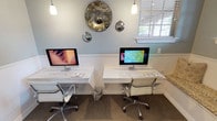 This business center is a thoughtfully designed single-floor workspace that combines functionality with refined aesthetic appeal. The room is finished in soft blue-gray tones with classic white wainscoting running along the lower portion of the walls, complemented by crown molding that adds an elegant architectural touch.

Two dedicated computer workstations are positioned along a continuous white desk surface, each equipped with an iMac, keyboard, and accessories. Ergonomic rolling chairs provide comfortable seating at each station. Pendant lighting hangs overhead, casting focused illumination across the workspace. A decorative arrangement of round mirrors, including an antique-style mercury glass piece flanked by smaller convex mirrors, serves as a striking focal point on the main wall.

Natural light enters through multiple windows fitted with white horizontal blinds, creating a bright and welcoming atmosphere. A French door with multi-pane glass panels leads to an adjacent area, maintaining an open visual connection. Built-in white shelving units with open cubbies provide storage and display space, and a printer station is conveniently integrated into the built-ins. A cushioned window bench adds a comfortable seating option, completing this polished and practical workspace.