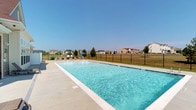 This community amenity space is highlighted by a generously sized outdoor swimming pool that serves as its central feature. The pool is filled with clear, blue water and is surrounded by a clean, light-colored concrete deck that provides ample space for lounging and relaxation. Poolside seating, including lounge chairs and a shaded umbrella area, offers residents a comfortable place to unwind under open skies.

The pool area is fully enclosed by a sturdy black iron fence, ensuring both safety and a defined boundary for the space. Beyond the fence, well-maintained green lawns stretch outward, framed by a backdrop of a charming residential neighborhood with a variety of single-family homes visible in the distance.

The setting benefits greatly from its open-air design, allowing for abundant natural light and sweeping views of the surrounding community under a wide, clear blue sky. The overall atmosphere is clean, bright, and inviting, making it an ideal gathering spot for outdoor recreation. The combination of the expansive pool, thoughtful deck layout, and picturesque neighborhood surroundings creates a welcoming and functional communal environment suited for leisure and social activity.