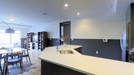 This single-floor space showcases a clean, modern aesthetic with an open-concept design that encourages a natural flow between the kitchen and living areas. The kitchen is a standout feature, anchored by a large island with crisp white countertops and an undermount stainless steel sink fitted with a contemporary arc faucet. Dark cabinetry on the island provides a striking contrast against the light surfaces, adding depth and sophistication to the space. The walls feature a two-tone treatment, with a bold dark lower half meeting a textured, light-toned upper section that adds visual interest and dimension throughout. Recessed lighting in the white ceiling ensures the space is well-lit and feels open. A framed doorway with dark trim leads to an adjacent area, enhancing the sense of connected, flowing space. The living area is visible in the background, furnished with open shelving units in a rich dark wood tone alongside white storage, offering both practicality and style. Light wood-toned flooring ties the entire space together, contributing to the warm yet contemporary ambiance that defines this thoughtfully designed interior.