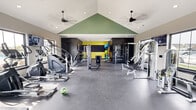 This fitness center offers a generously sized, single-level workout space designed to accommodate a comprehensive range of exercise activities. The room's most striking architectural feature is its vaulted ceiling, accented by a bold green triangular panel that rises toward the peak, creating a sense of height and visual interest. Two large ceiling fans are suspended from the vaulted ceiling, ensuring consistent airflow throughout the space. Recessed lighting supplements the natural light that enters through windows along the perimeter walls, contributing to a bright and energizing atmosphere.

The flooring throughout is durable rubber gym matting in a charcoal tone, providing cushioning and traction suited to high-impact activity. Along one side of the room, a row of treadmills and elliptical machines faces large windows, allowing users to exercise with an outdoor view. Strength training equipment, including cable machines and multi-functional weight stations, is distributed throughout the space. A dedicated free weights area features a full dumbbell rack, while a functional training corner houses kettlebells, resistance tools, and a medicine ball storage rack. Wall-mounted televisions and large mirrors enhance the overall training environment. The accent wall at the rear features a vivid yellow tone, adding a dynamic visual focal point.