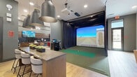 Located in Mesa, AZ, this single-floor amenity lounge offers a well-appointed, multi-functional gathering space designed for both relaxation and recreation. The room is thoughtfully divided into distinct zones, each contributing to the space's dynamic character.

The lounge area features light wood-look flooring, high ceilings, and floor-to-ceiling windows that invite natural light and provide views of the surrounding exterior. A bold feature wall clad in dark brick tile and warm wood paneling anchors a wall-mounted flat-screen TV, flanked by vibrant rust-orange barrel chairs and a sleek glass coffee table, creating an inviting social hub. A curated gallery wall of sports-themed artwork adds a spirited, energetic touch to an adjacent seating nook with a neutral sofa and accent chairs.

A separate lounge area showcases a textured, geometric-patterned white tile accent wall adorned with contemporary black-and-white abstract art. Plush blush swivel chairs and natural wood stump side tables lend a relaxed, organic elegance to this corner.

The standout feature of the space is a full indoor golf simulator, complete with a synthetic turf hitting mat, a large high-definition projection screen, ceiling-mounted projector, and professional club storage. Dark curtain panels frame the simulator bay, creating an immersive experience. A counter-height bar with pendant lighting serves as a transitional element between the simulator and lounge zones, making this space ideal for social and recreational use.