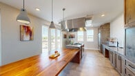 Located at 1236 Water Tower Circle in Greenville, SC, this thoughtfully designed kitchen delivers a refined blend of professional functionality and warm, contemporary aesthetics. The space is anchored by a striking long butcher block island that serves as both a prep surface and a gathering point, illuminated overhead by a trio of modern pendant lights that add an industrial yet inviting character.

At the heart of the cooking area, a high-performance gas range sits beneath a bold stainless steel range hood, which commands attention as a central architectural element. Flanking the range are rich, warm-toned wood cabinetry units that extend floor to ceiling, providing generous storage. A built-in stainless steel refrigerator integrates seamlessly into the cabinetry wall, lending a cohesive, upscale appearance.

The countertops throughout feature a smooth, light-toned stone surface, offering ample workspace along the perimeter. A deep undermount sink with a matte black faucet adds a refined, modern contrast to the surrounding finishes. Light stone-tiled flooring runs throughout, contributing to the bright, open feel of the room.

Multiple windows along the exterior walls invite abundant natural light and offer views of the surrounding Greenville landscape. Recessed ceiling lighting supplements the natural illumination, ensuring the space is well-lit throughout the day.
