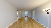 Located at 134 College Hwy in Southampton, Massachusetts, this single-floor unit offers a straightforward and functional layout with a clean, neutral design throughout. Natural wood-toned flooring extends across the main living areas, lending warmth and continuity to the space. The open living area is generously proportioned, with light gray walls, white trim, and baseboard heating that runs along the perimeter. A wall-mounted air conditioning unit provides climate control in one of the rooms, which also benefits from a large window that allows natural light to fill the space. The kitchen is efficiently designed with white cabinetry, tile backsplash, laminate countertops, a stainless steel sink, and white appliances including an electric range with overhead ventilation. A window above the sink brings in additional natural light. The bedroom features light wood flooring, a built-in closet with sliding doors, and direct access to the rest of the unit. One room showcases a distinctive tiled accent wall with a subtle grid pattern, adding a unique textural element to the interior. The full bathroom is finished with neutral tile flooring, a white vanity with storage, a toilet, and a tub-shower combination, all set within a bright, well-lit space. Overall, the unit presents a tidy and move-in ready environment.