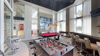This contemporary amenity space delivers a sophisticated and energetic atmosphere designed for entertainment and social interaction. The centerpiece of the room is a bold red felt pool table, perfectly positioned beneath a striking layered pendant light fixture that adds a touch of elegance to the space. Adjacent to it, a foosball table invites casual competition, while a large multi-panel video wall mounted on a sleek glass partition serves as a dynamic focal point. The soaring double-height ceilings, accented by a dramatic geometric black ceiling treatment, create an impressive sense of volume and grandeur. Floor-to-ceiling windows and sheer white drapery line the walls, flooding the interior with abundant natural light and offering views of lush outdoor greenery. A patterned area rug in muted tones grounds the space, complementing the modern bar-height seating along the perimeter. An adjacent lounge area, partially visible through the glass partition, features plush white furnishings, providing a more relaxed retreat. The overall design seamlessly blends high-end finishes with a playful, resort-style character, making it an ideal gathering space for residents or guests.