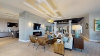 This elegantly designed single-floor clubhouse offers a refined communal experience through a series of thoughtfully curated spaces. The centerpiece is a spacious main lounge anchored by a sleek linear fireplace set into a textured stone-clad accent wall, above which a large flat-screen TV is mounted. Coffered ceilings with crisp white beams lend architectural distinction overhead, while a glamorous gold chandelier adds a striking focal point. Seating is arranged in relaxed groupings featuring woven rattan chairs, upholstered sofas, and warm wood side tables, creating an inviting and stylish atmosphere.

Elegant columns define the transition to additional seating zones, where a baby grand piano and curated artwork contribute to a cultured ambiance. Natural light pours in through large windows fitted with charcoal drape panels, offering views of palm trees and the surrounding outdoor amenities.

A dedicated beverage bar is outfitted with dark granite countertops, light wood cabinetry, a sink, coffee station, and wall-mounted screens — ideal for casual gatherings or productive mornings. Adjacent to this is a conference-style room featuring a long communal table, pendant lighting, a fireplace, bold artwork, and direct access to the outdoors. Quiet reading alcoves with plush accent chairs complete this well-appointed space.