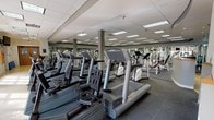 This fitness facility is organized across a single level, offering a comprehensive workout environment designed to accommodate a wide range of exercise needs. Guests are welcomed through a distinctive entrance lobby featuring a bold, curvilinear floor pattern in contrasting tones of tan, blue, and green, paired with a rounded reception desk beneath a circular ceiling detail — creating an inviting first impression.

The main workout floor is expansive, with a drop ceiling fitted with recessed and panel lighting that evenly illuminates the space throughout. The cardio zone houses rows of treadmills, ellipticals, stair climbers, and stationary bikes. Natural light filters in through a bank of windows along one wall, lending an open, airy quality to this area, while mounted televisions provide additional engagement for users.

The strength training section is well-stocked with a broad selection of selectorized machines, cable stations, power racks, squat racks, and benches. A dedicated free weights area features a full dumbbell rack alongside multiple adjustable benches. Floor-to-ceiling mirrors line the walls, enhancing the sense of depth and allowing users to monitor their form. The rubber flooring throughout the workout areas provides a durable, functional surface, while instructional posters and informational signage are displayed across the walls.