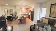 This single-floor residence offers an open-concept layout that seamlessly connects the kitchen, dining, and living areas, creating a bright and functional living environment. The kitchen is a standout feature, showcasing warm medium-toned wood cabinetry, dark granite countertops, and a suite of black appliances including a refrigerator, oven, and built-in microwave. A central kitchen island with seating provides a convenient space for casual dining or meal preparation, while pendant lighting above adds a welcoming ambiance. The kitchen flows naturally into a dedicated dining area, which benefits from an abundance of natural light and offers ample space for a full dining set. Adjacent to the kitchen, the living area features plush carpeting and a cozy, well-furnished layout, anchored by a glass-top coffee table and comfortable seating. Hardwood-style flooring in the kitchen and dining zones transitions smoothly into the carpeted living space, adding visual warmth throughout. A large sliding door or window at the edge of the living area brings in generous natural light and hints at outdoor access. The overall ambiance is warm, cohesive, and thoughtfully designed for comfortable everyday living.
