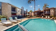 Located at 22221 Cypresswood Dr in Spring, Texas, this outdoor amenity area serves as a central gathering space within a residential community. The focal point is a generously sized swimming pool with clear, turquoise water, complemented by a separate spa enclosure, both enclosed within a wrought iron fence for safety and privacy.

The sun deck surrounding the pool is finished with a textured concrete surface and outfitted with multiple lounging chairs and umbrella-shaded seating areas, providing ample space for relaxation and outdoor enjoyment. Decorative brick planters with colorful tropical plants and manicured shrubs add a welcoming, resort-like ambiance to the space.

The surrounding residential buildings feature a blend of brick and wood-panel exterior finishes, with multi-level balconies and prominent chimneys that contribute to a classic architectural character. Tall pine trees frame the perimeter of the space, offering natural shade and a sense of privacy. Well-maintained walkways and lush green landscaping connect the pool area to the surrounding buildings, creating a cohesive and inviting outdoor environment. Under the wide Texas sky, this amenity space offers a bright, open, and comfortable retreat for residents throughout the year.