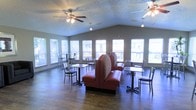 This inviting single-floor space functions as a communal dining or lounge area, thoughtfully designed to accommodate both casual gatherings and relaxed seating. The room's most notable architectural feature is its vaulted ceiling, which adds a sense of openness and volume, complemented by a textured finish overhead. Multiple ceiling fans are suspended throughout, ensuring comfortable airflow across the space.

Natural light is a defining characteristic, with a generous row of large windows lining the walls on multiple sides, fitted with horizontal blinds for adjustable light control. The abundance of windows creates a bright, airy atmosphere while offering views to the surrounding exterior.

The flooring consists of dark hardwood-style planks, providing a warm contrast to the light gray walls and bright ceiling. Seating is varied and practical, featuring centrally placed red upholstered booth seating with high backs as a focal point, surrounded by multiple café-style bistro tables paired with metal chairs arranged along the windowed perimeter. The layout encourages both intimate conversations and larger group interactions, making the space versatile and welcoming for everyday use.