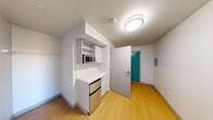 Located at 1815 Bellevue Ave in Seattle's Capitol Hill neighborhood, Unit 207 is a thoughtfully designed single-floor studio. The unit features warm hardwood-style flooring throughout, clean white walls, and a bright, modern aesthetic that maximizes the efficiency of the space.

The main living area is open and versatile, benefiting from natural light that filters in through a tall, narrow window alongside a larger operable window, offering views of the surrounding urban streetscape. The clean, uninterrupted wall space provides flexibility for furnishing and personalization.

A built-in kitchenette is neatly integrated into one wall, featuring upper and lower white cabinetry, a microwave, a compact stainless steel refrigerator, and a white countertop workspace. A wall-mounted utility panel is also present near the entry. The distinctive teal entry door adds a pop of color and opens to a small foyer area with ceiling-mounted lighting.

The bathroom is clean and functional, finished with the same light flooring as the rest of the unit. It includes a walk-in shower with a rod, a toilet, a wall-mounted cabinet, and a vanity sink, all finished in white for a cohesive, minimal look.

Overall, Unit 207 presents a clean, modern, and efficiently organized living space well-suited for urban living in Seattle.
